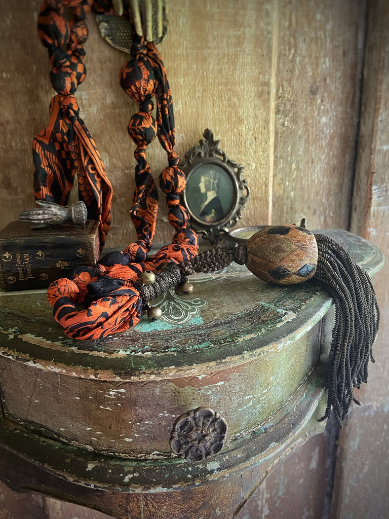 Aesthetic Movement inspired long silk sautoir necklace with antique tassel embellishment. Bohemian style from vintage textiles by Pavilion Parade. Decorative objects including a woven item and a small portrait on a wooden surface.